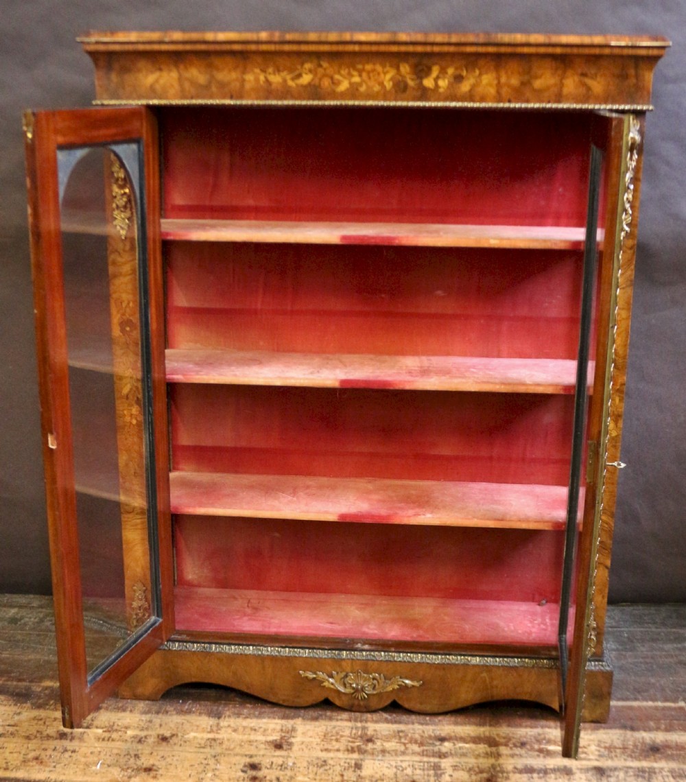 A Victorian burr walnut pier cabinet featuring floral marquetry inlays, gilt ormolu mounts, and double arched glass doors revealing shelves lined in red fabric. View II