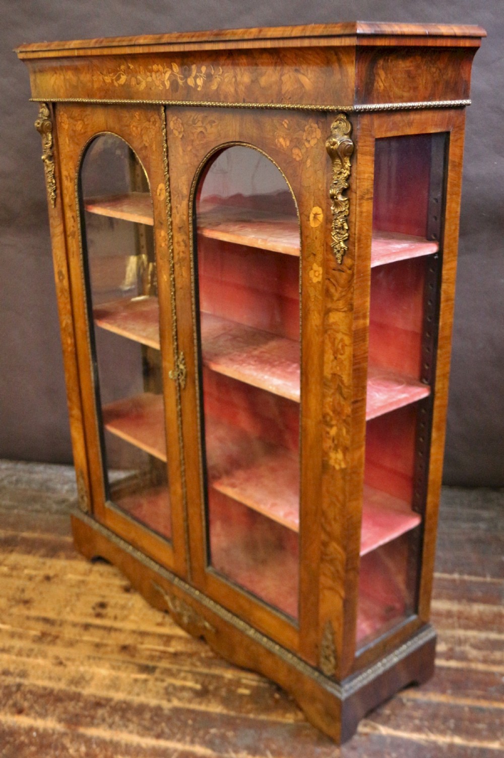 A Victorian burr walnut pier cabinet featuring floral marquetry inlays, gilt ormolu mounts, and double arched glass doors revealing shelves lined in red fabric. View III