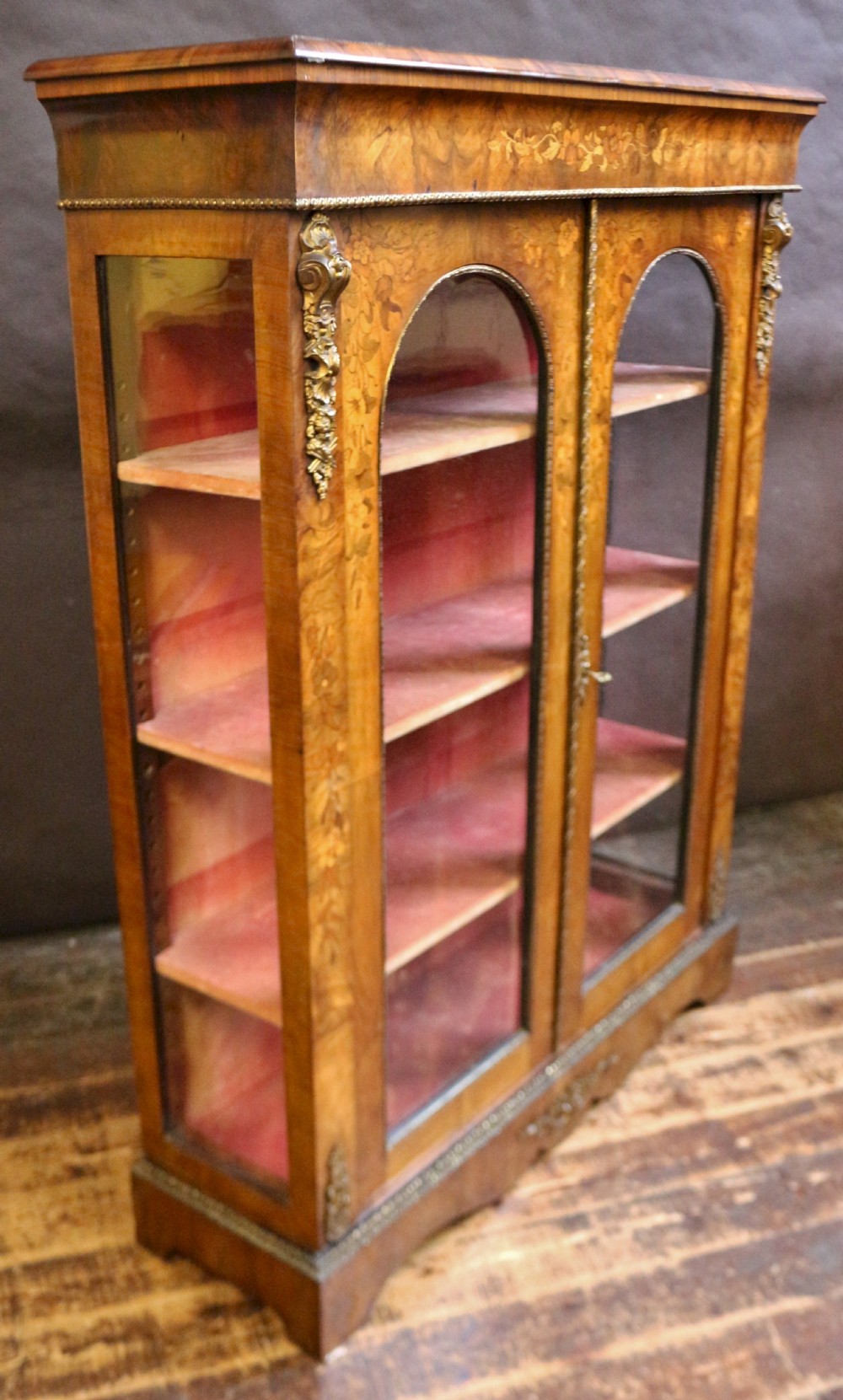 A Victorian burr walnut pier cabinet featuring floral marquetry inlays, gilt ormolu mounts, and double arched glass doors revealing shelves lined in red fabric. View IV