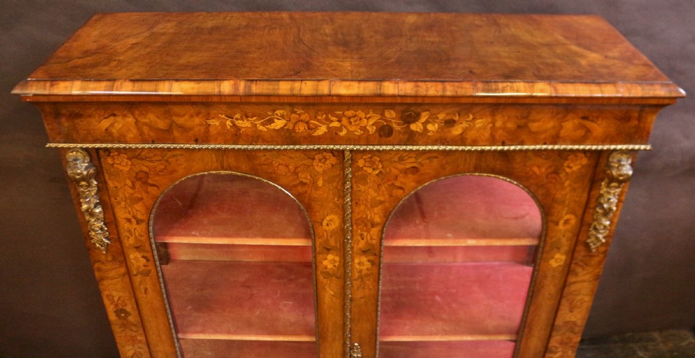 A Victorian burr walnut pier cabinet featuring floral marquetry inlays, gilt ormolu mounts, and double arched glass doors revealing shelves lined in red fabric. View V