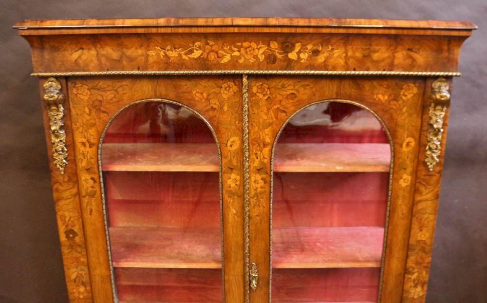 A Victorian burr walnut pier cabinet featuring floral marquetry inlays, gilt ormolu mounts, and double arched glass doors revealing shelves lined in red fabric. View VI