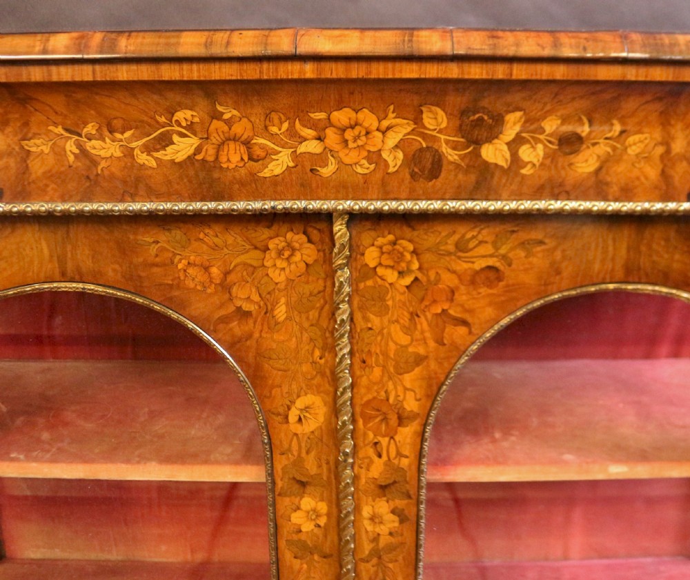 A Victorian burr walnut pier cabinet featuring floral marquetry inlays, gilt ormolu mounts, and double arched glass doors revealing shelves lined in red fabric. View VII