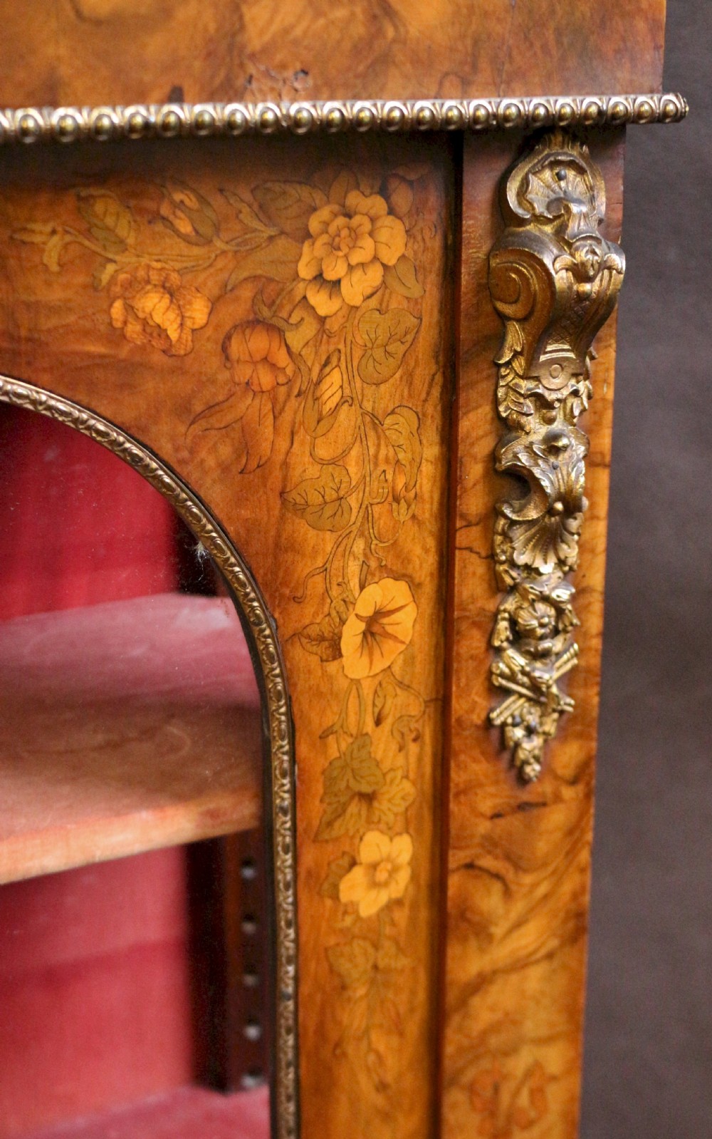A Victorian burr walnut pier cabinet featuring floral marquetry inlays, gilt ormolu mounts, and double arched glass doors revealing shelves lined in red fabric. View IX