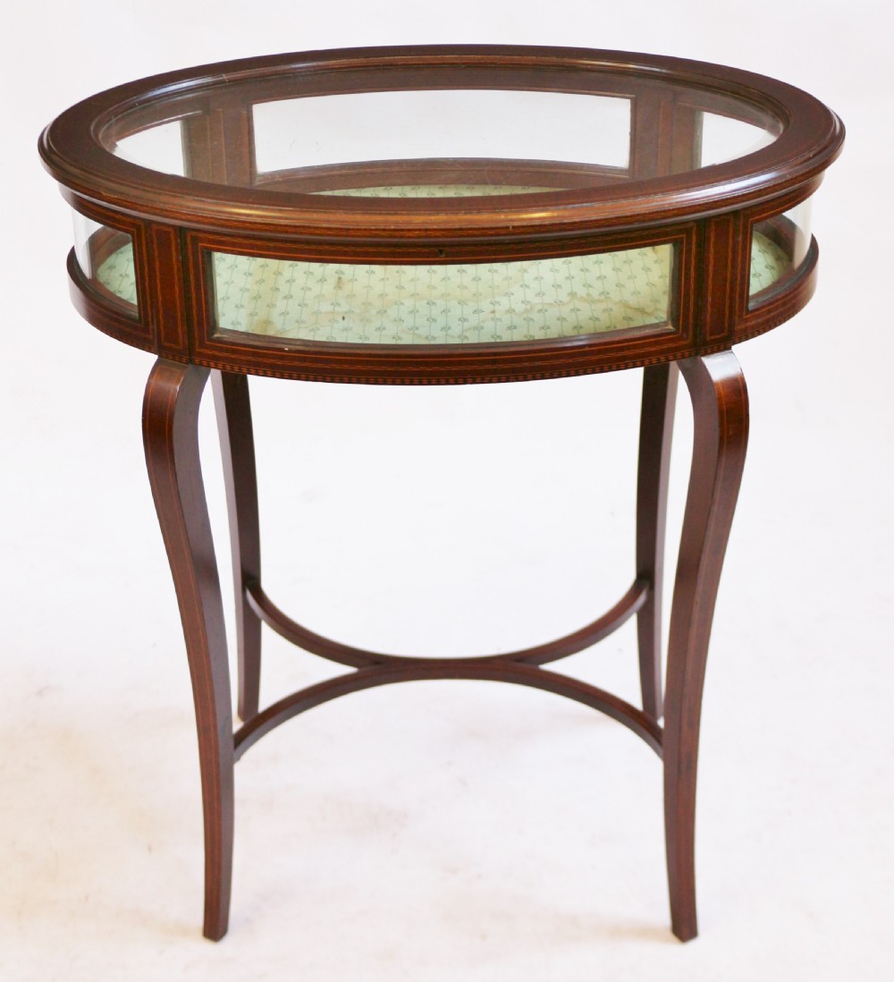 An Edwardian oval mahogany bijouterie table with glass top and sides, satinwood line inlay, curved legs, and an X-form stretcher.
