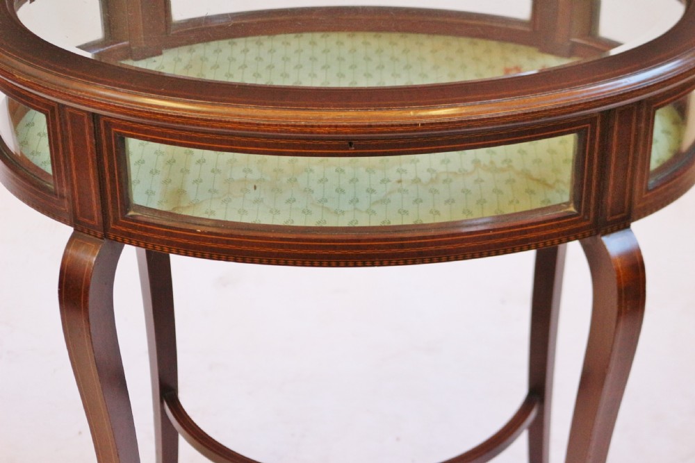 An Edwardian oval mahogany bijouterie table with glass top and sides, satinwood line inlay, curved legs, and an X-form stretcher. View VII