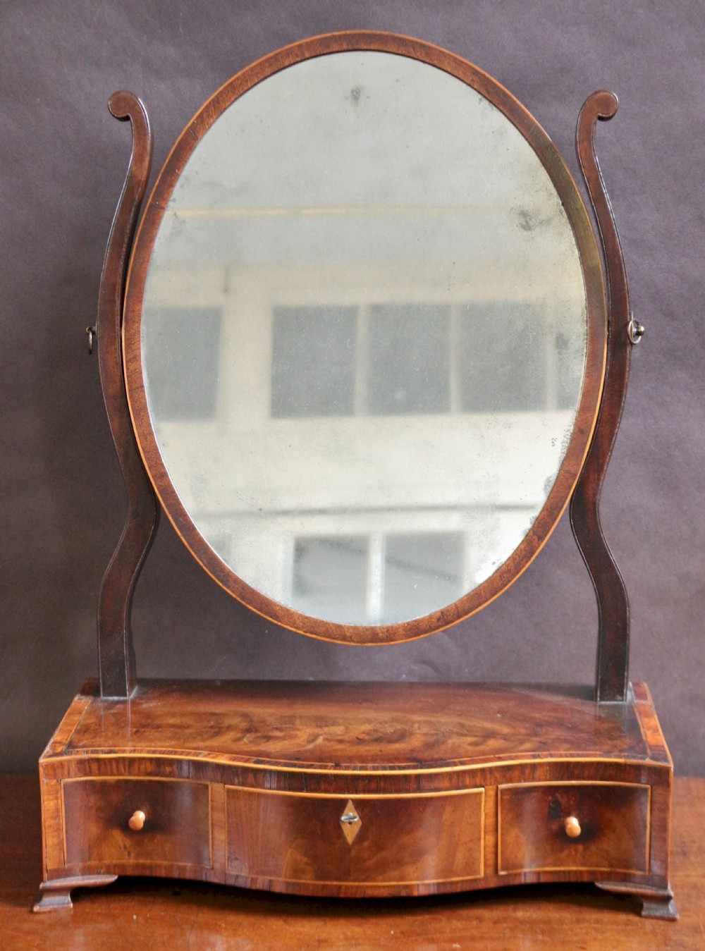 A George III mahogany toilet mirror featuring an oval adjustable mirror plate mounted on curved uprights above a serpentine base with three small drawers.