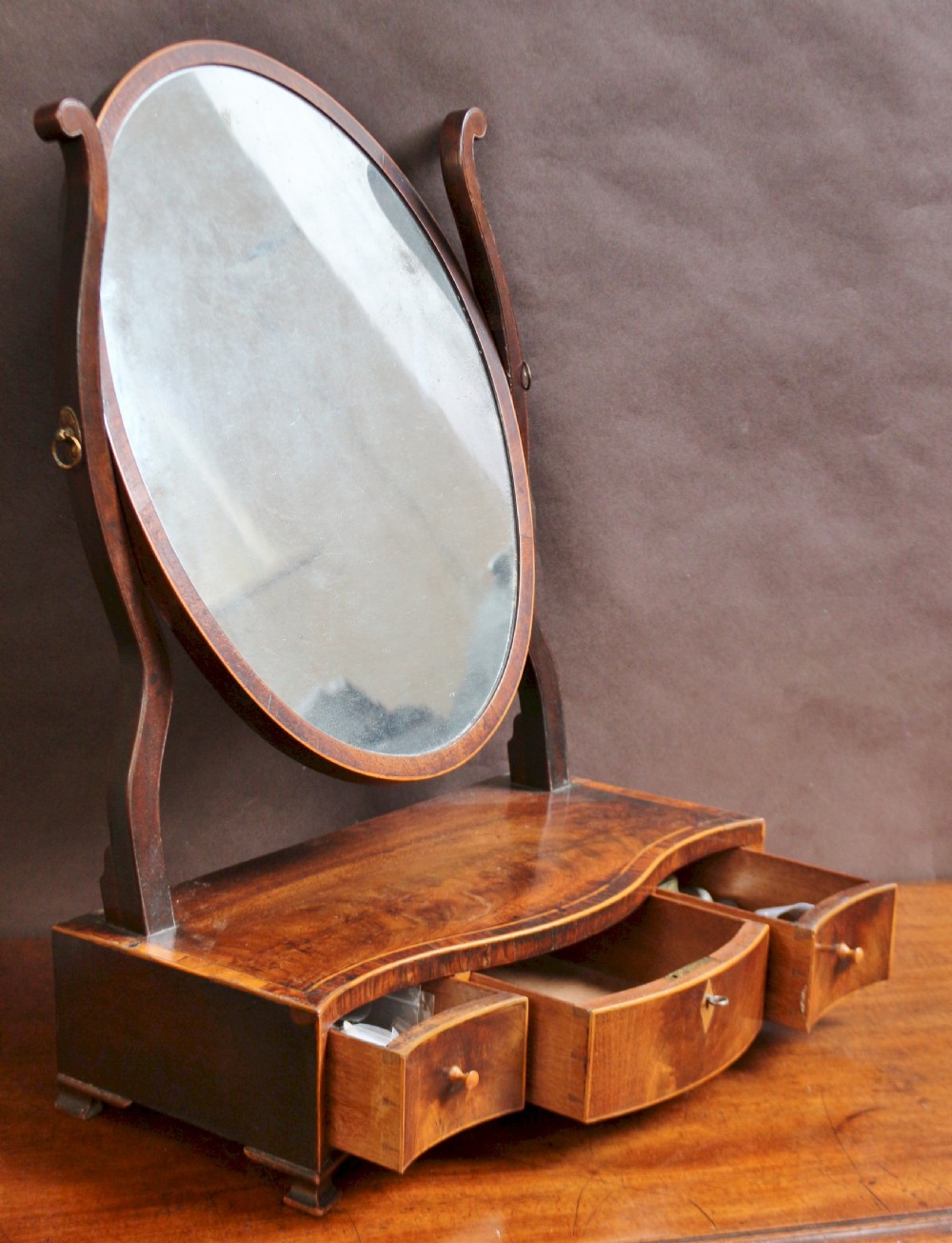 A George III mahogany toilet mirror featuring an oval adjustable mirror plate mounted on curved uprights above a serpentine base with three small drawers. View II