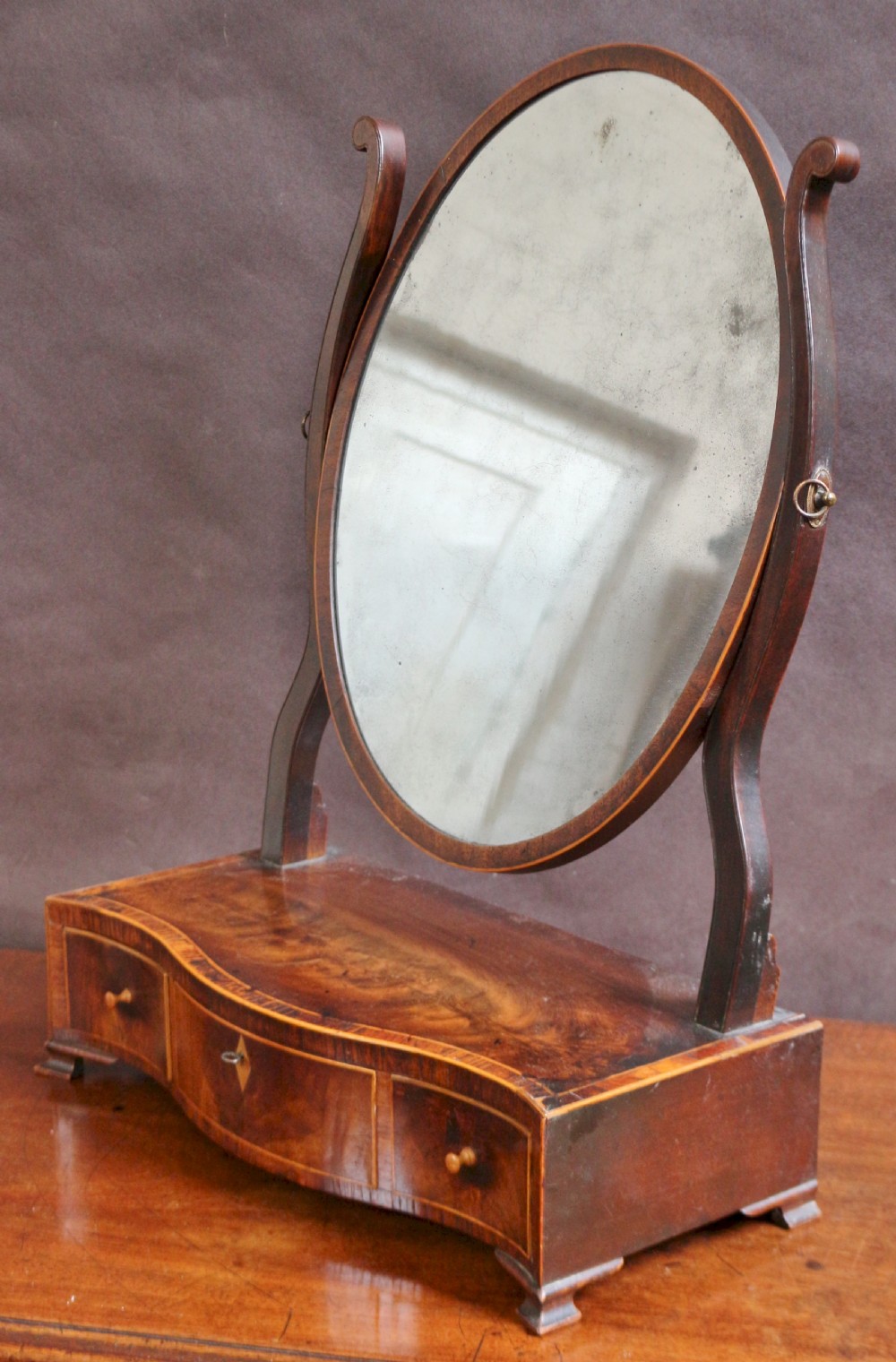 A George III mahogany toilet mirror featuring an oval adjustable mirror plate mounted on curved uprights above a serpentine base with three small drawers. View III