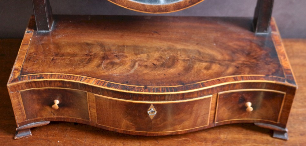 A George III mahogany toilet mirror featuring an oval adjustable mirror plate mounted on curved uprights above a serpentine base with three small drawers. View IV