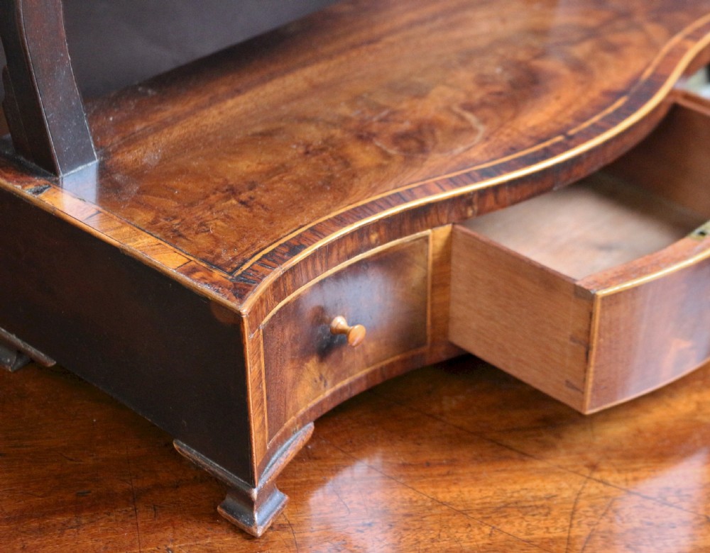 A George III mahogany toilet mirror featuring an oval adjustable mirror plate mounted on curved uprights above a serpentine base with three small drawers. View V