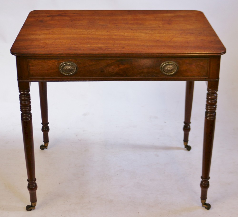 A George IV mahogany side table featuring a single frieze drawer with brass ring handles, turned legs, and brass castors.