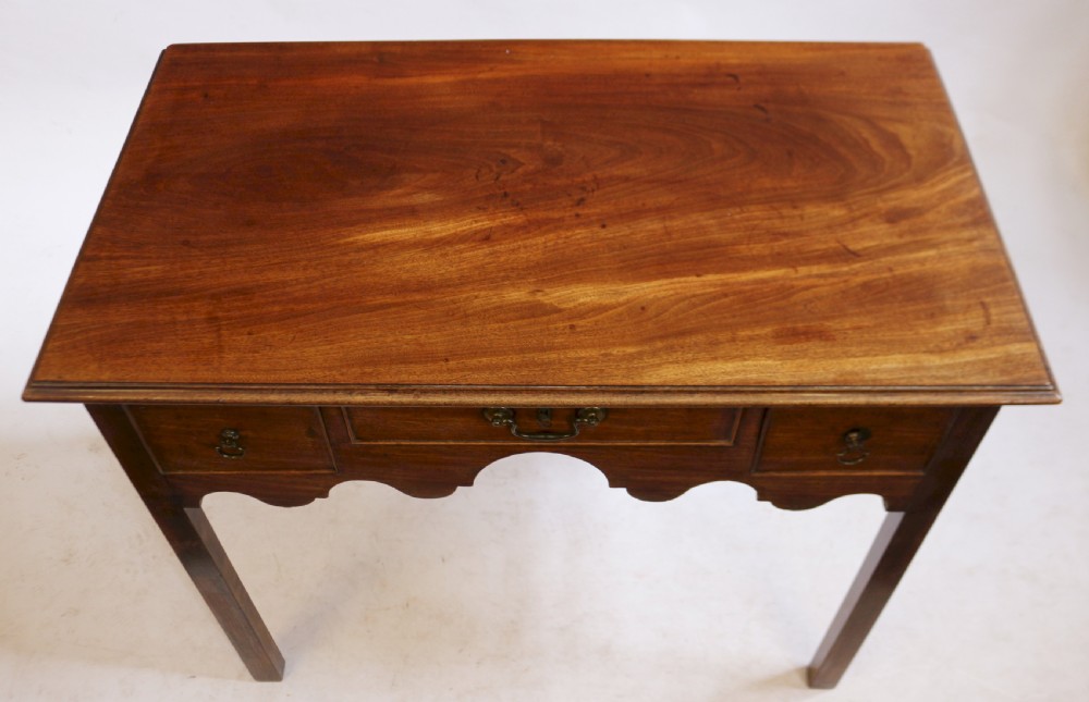 A George III mahogany lowboy table featuring a moulded top over three drawers with brass handles and a scalloped apron on square legs. View IV