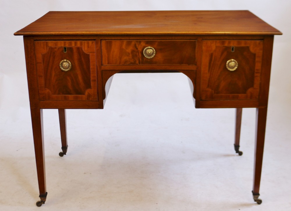 A George III mahogany kneehole desk featuring three inlaid drawers, central kneehole arch, brass handles, and square tapered legs with brass castors.