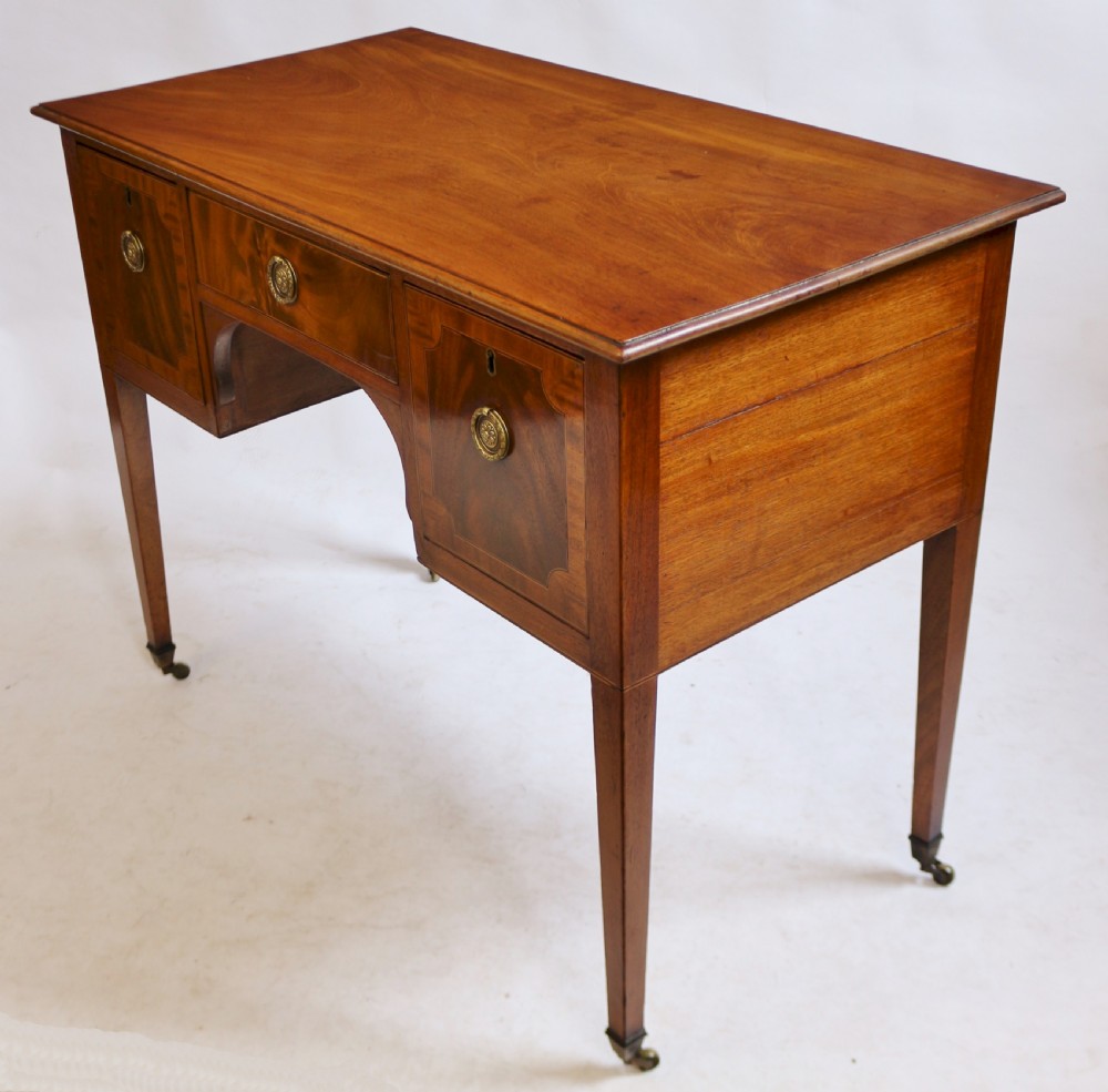 A George III mahogany kneehole desk featuring three inlaid drawers, central kneehole arch, brass handles, and square tapered legs with brass castors. View III