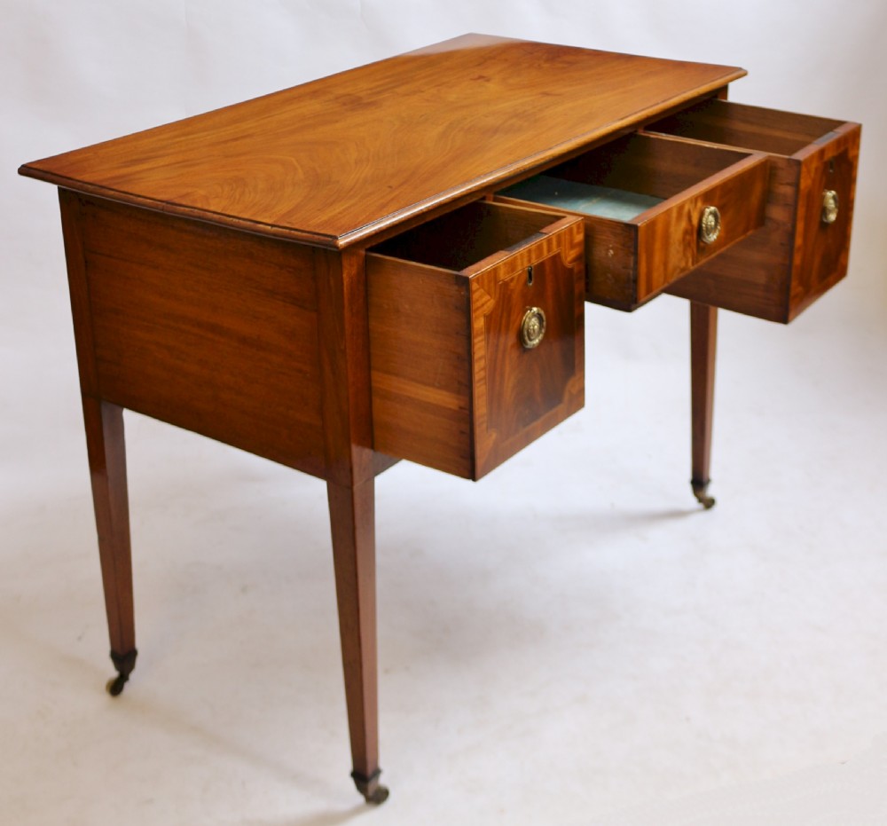 A George III mahogany kneehole desk featuring three inlaid drawers, central kneehole arch, brass handles, and square tapered legs with brass castors. View IV