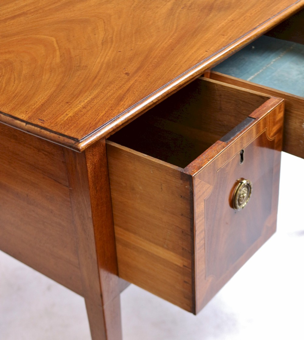 A George III mahogany kneehole desk featuring three inlaid drawers, central kneehole arch, brass handles, and square tapered legs with brass castors. View V