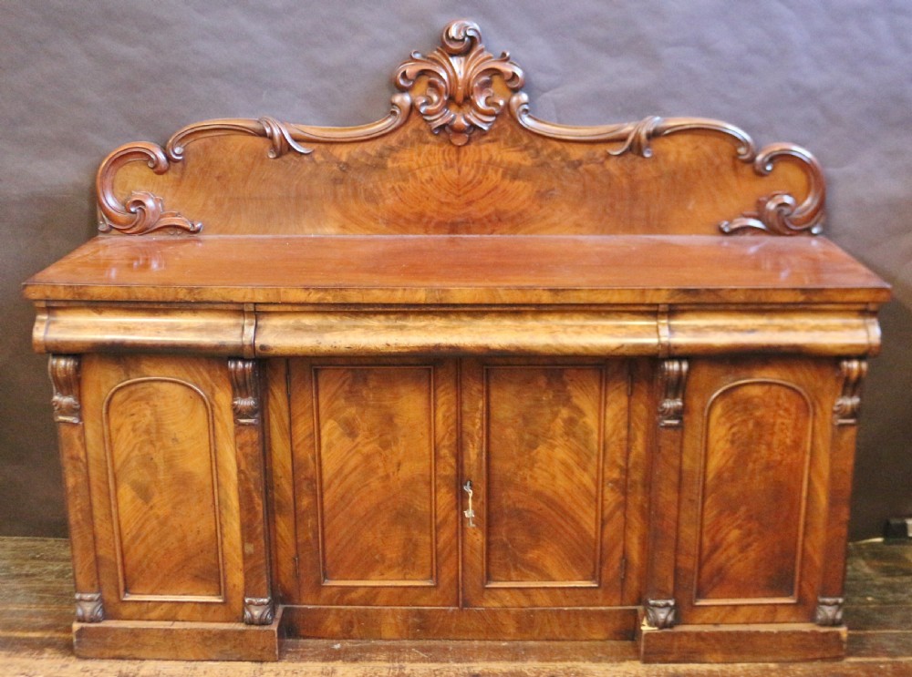 A Victorian mahogany four-door sideboard featuring a scroll-carved backboard, flame mahogany doors, and a solid plinth base.