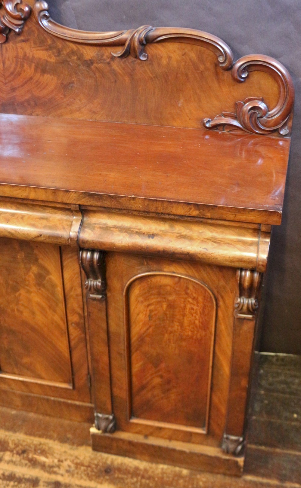 A Victorian mahogany four-door sideboard featuring a scroll-carved backboard, flame mahogany doors, and a solid plinth base. View V
