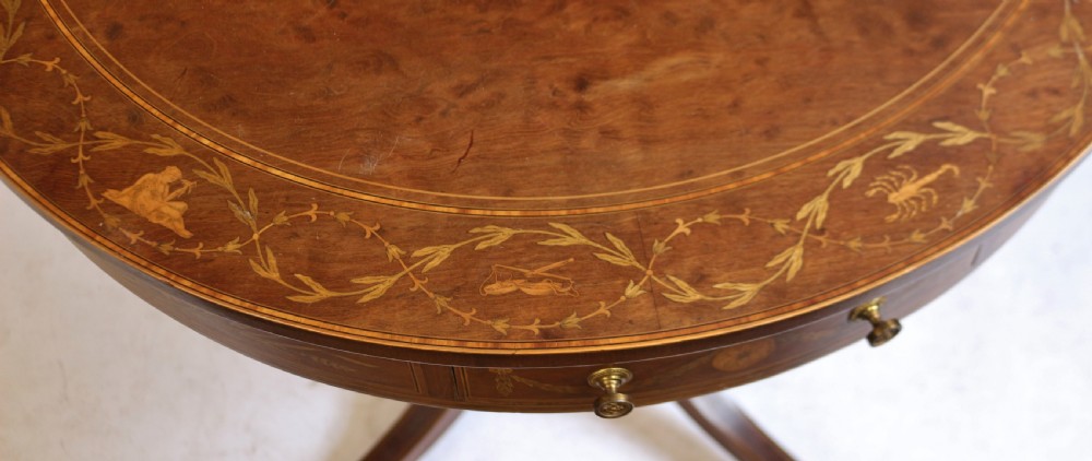 An Edwardian mahogany circular drum table featuring floral and zodiac marquetry inlays, a frieze drawer with dual brass knobs, and a central pedestal resting on four splayed legs with brass castors. View VI