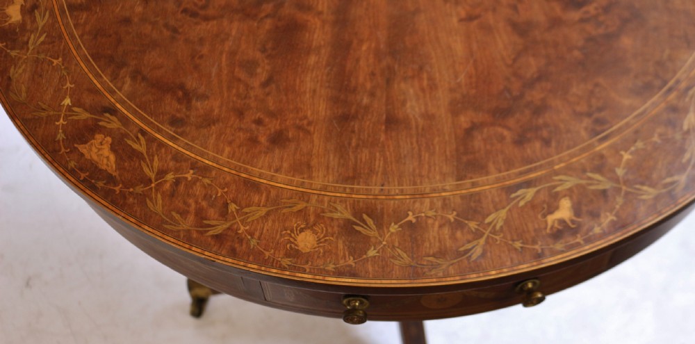 An Edwardian mahogany circular drum table featuring floral and zodiac marquetry inlays, a frieze drawer with dual brass knobs, and a central pedestal resting on four splayed legs with brass castors. View VII