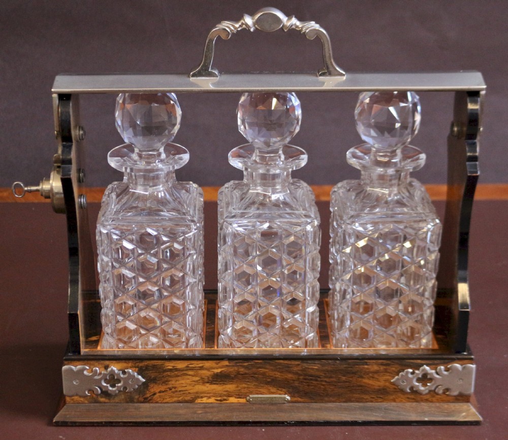 A late Victorian three-bottle tantalus constructed from dark figured coromandel wood with silver-plated corner mounts and three square cut-crystal decanters with faceted stoppers.