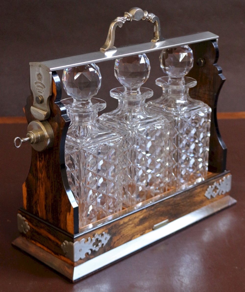 A late Victorian three-bottle tantalus constructed from dark figured coromandel wood with silver-plated corner mounts and three square cut-crystal decanters with faceted stoppers. View II