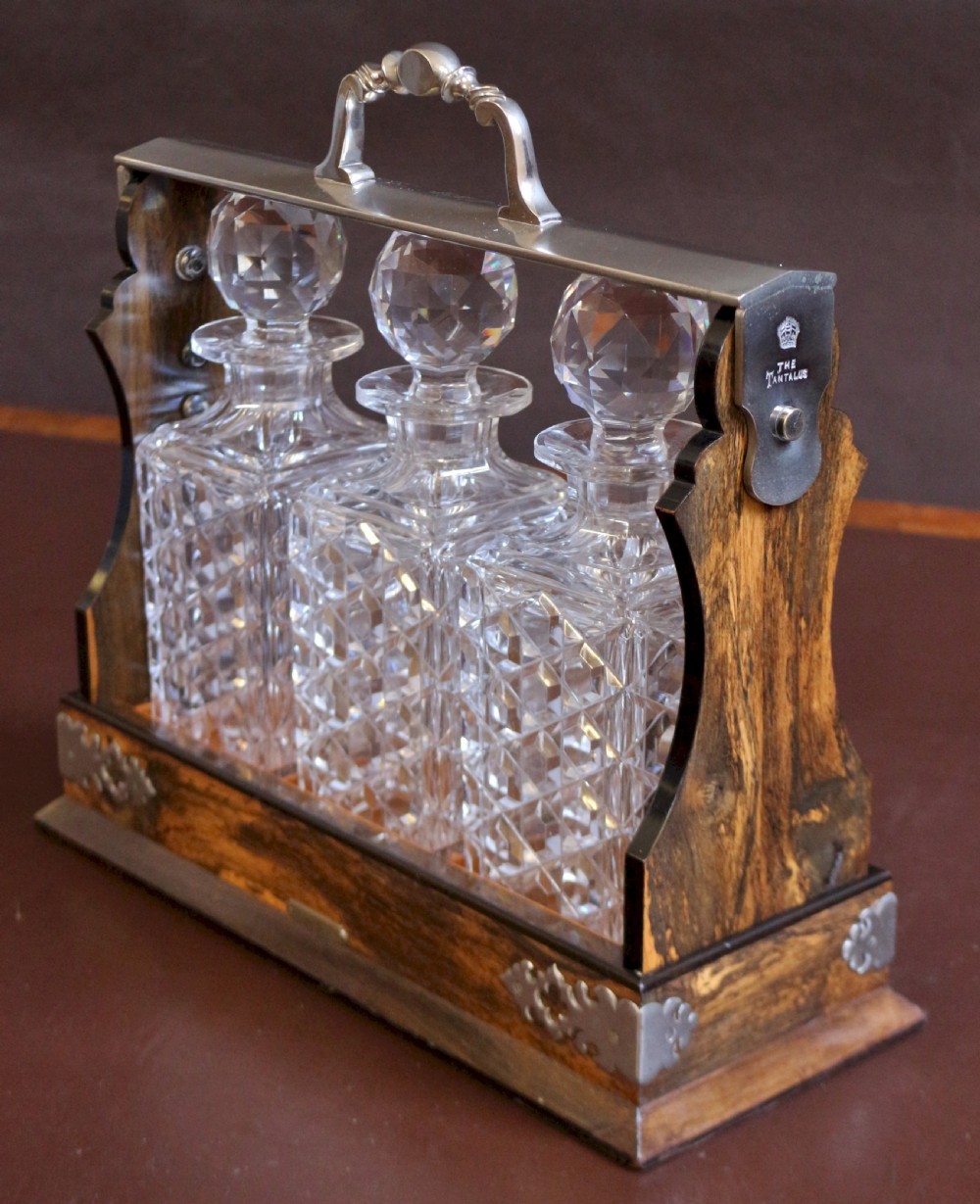 A late Victorian three-bottle tantalus constructed from dark figured coromandel wood with silver-plated corner mounts and three square cut-crystal decanters with faceted stoppers. View III