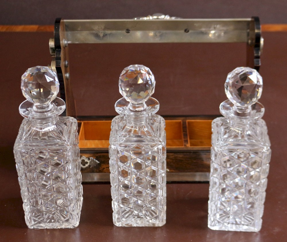 A late Victorian three-bottle tantalus constructed from dark figured coromandel wood with silver-plated corner mounts and three square cut-crystal decanters with faceted stoppers. View VIII