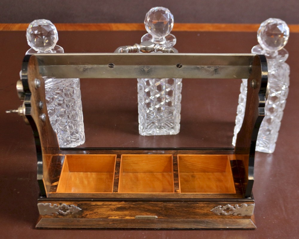 A late Victorian three-bottle tantalus constructed from dark figured coromandel wood with silver-plated corner mounts and three square cut-crystal decanters with faceted stoppers. View IX