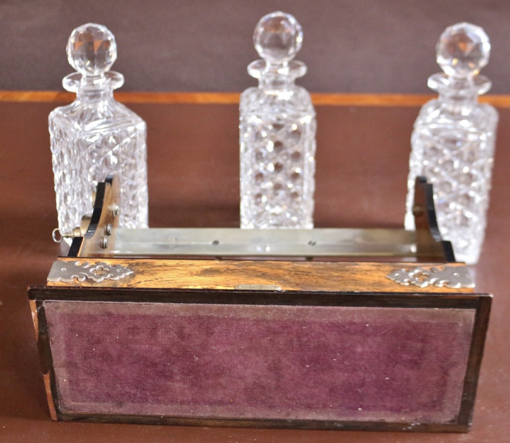 A late Victorian three-bottle tantalus constructed from dark figured coromandel wood with silver-plated corner mounts and three square cut-crystal decanters with faceted stoppers. View X