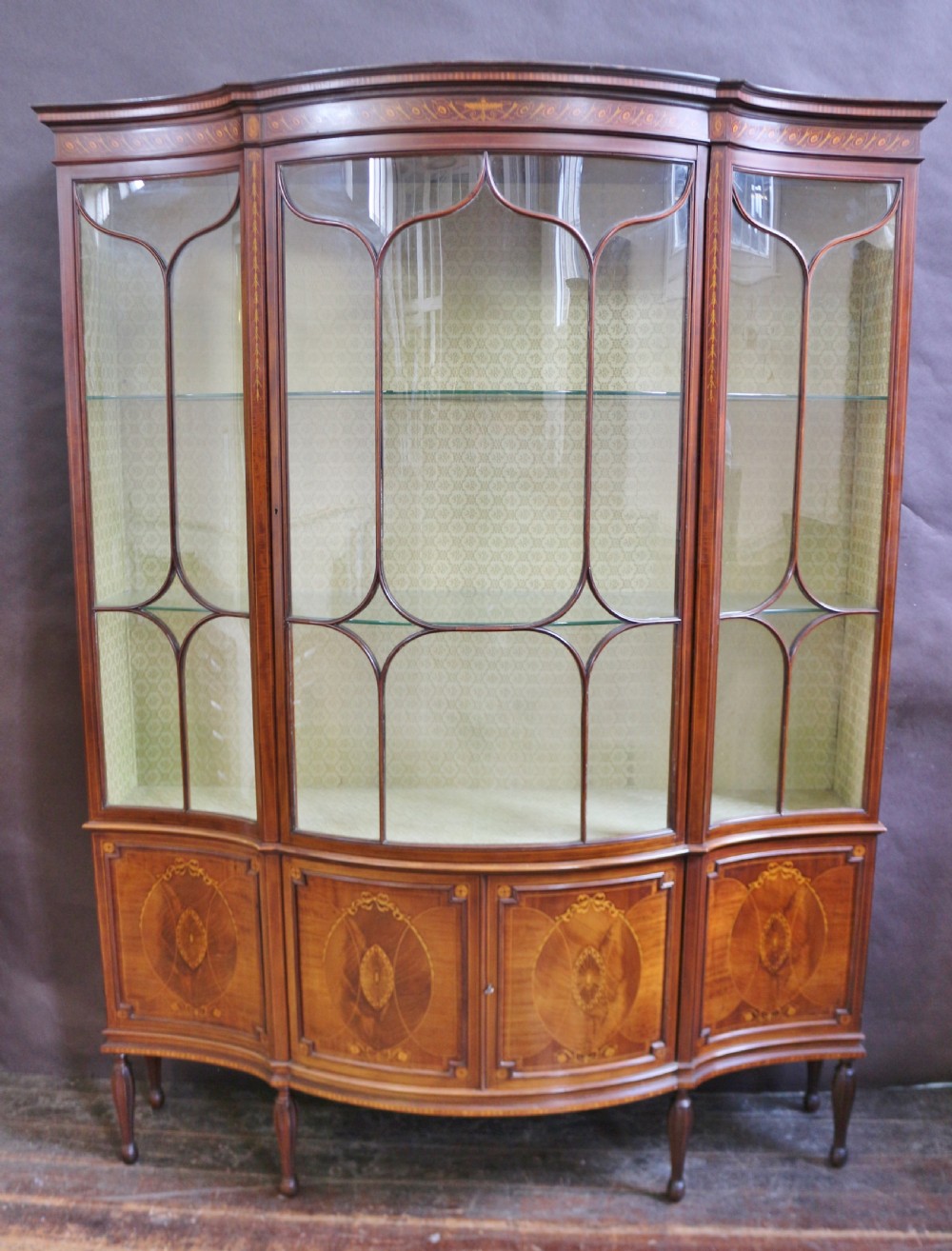 A late 19th-century mahogany display cabinet featuring three glazed doors with curved astragal glazing bars above inlaid lower panels and turned legs.