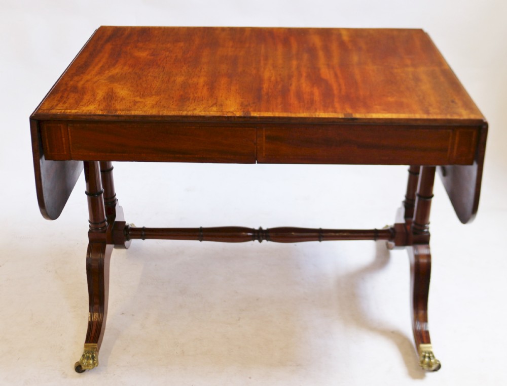 A Regency mahogany sofa table featuring drop-leaf ends, satinwood crossbanding, two frieze drawers, and splayed legs terminating in brass paw castors.