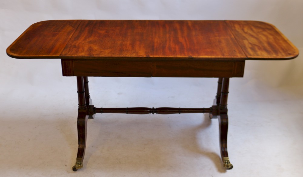 A Regency mahogany sofa table featuring drop-leaf ends, satinwood crossbanding, two frieze drawers, and splayed legs terminating in brass paw castors. View II