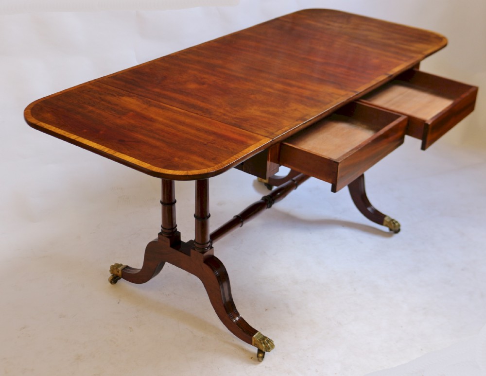 A Regency mahogany sofa table featuring drop-leaf ends, satinwood crossbanding, two frieze drawers, and splayed legs terminating in brass paw castors. View III