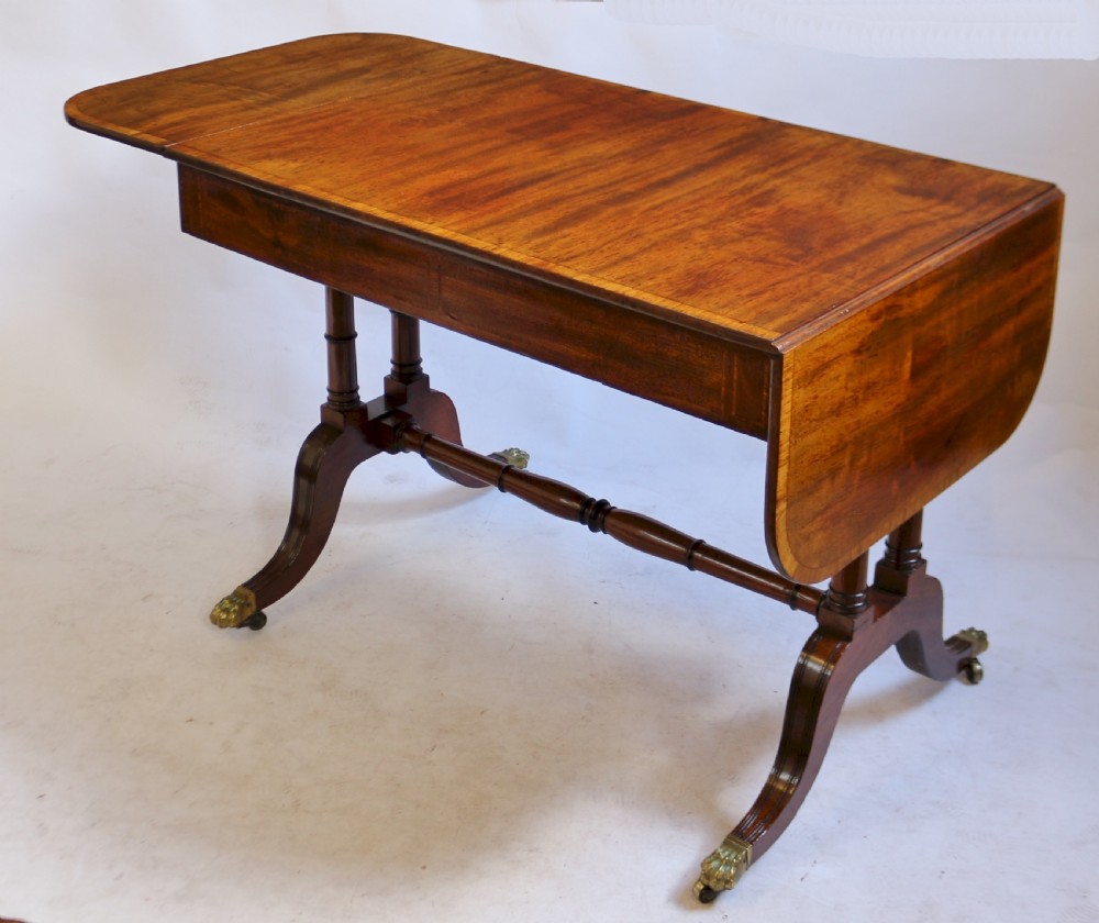 A Regency mahogany sofa table featuring drop-leaf ends, satinwood crossbanding, two frieze drawers, and splayed legs terminating in brass paw castors. View IV