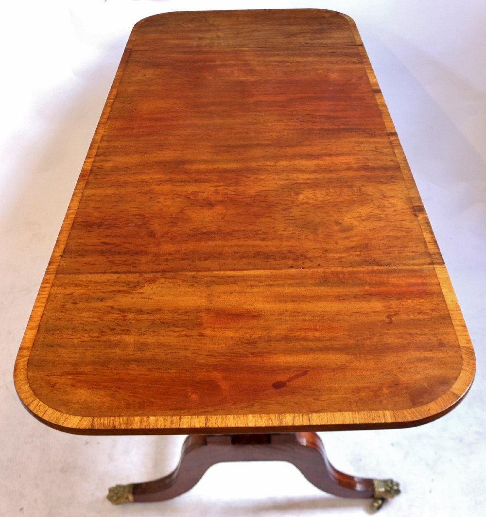 A Regency mahogany sofa table featuring drop-leaf ends, satinwood crossbanding, two frieze drawers, and splayed legs terminating in brass paw castors. View V