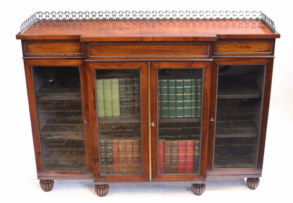 An antique Regency rosewood breakfront side cabinet featuring four glazed doors, a brass gallery rail along the top, and turned fluted feet.