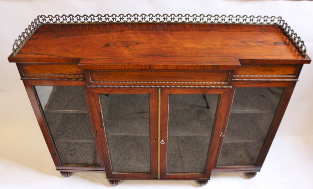 An antique Regency rosewood breakfront side cabinet featuring four glazed doors, a brass gallery rail along the top, and turned fluted feet. View V