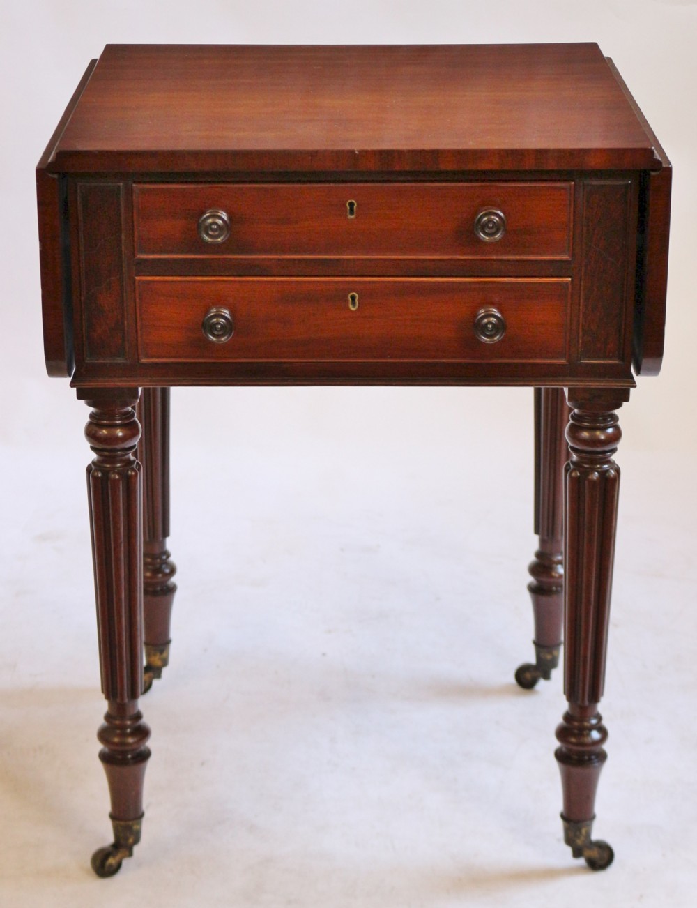A William IV mahogany drop-leaf side table featuring two drawers with turned knobs, brass keyhole escutcheons, and reeded legs on brass castors.