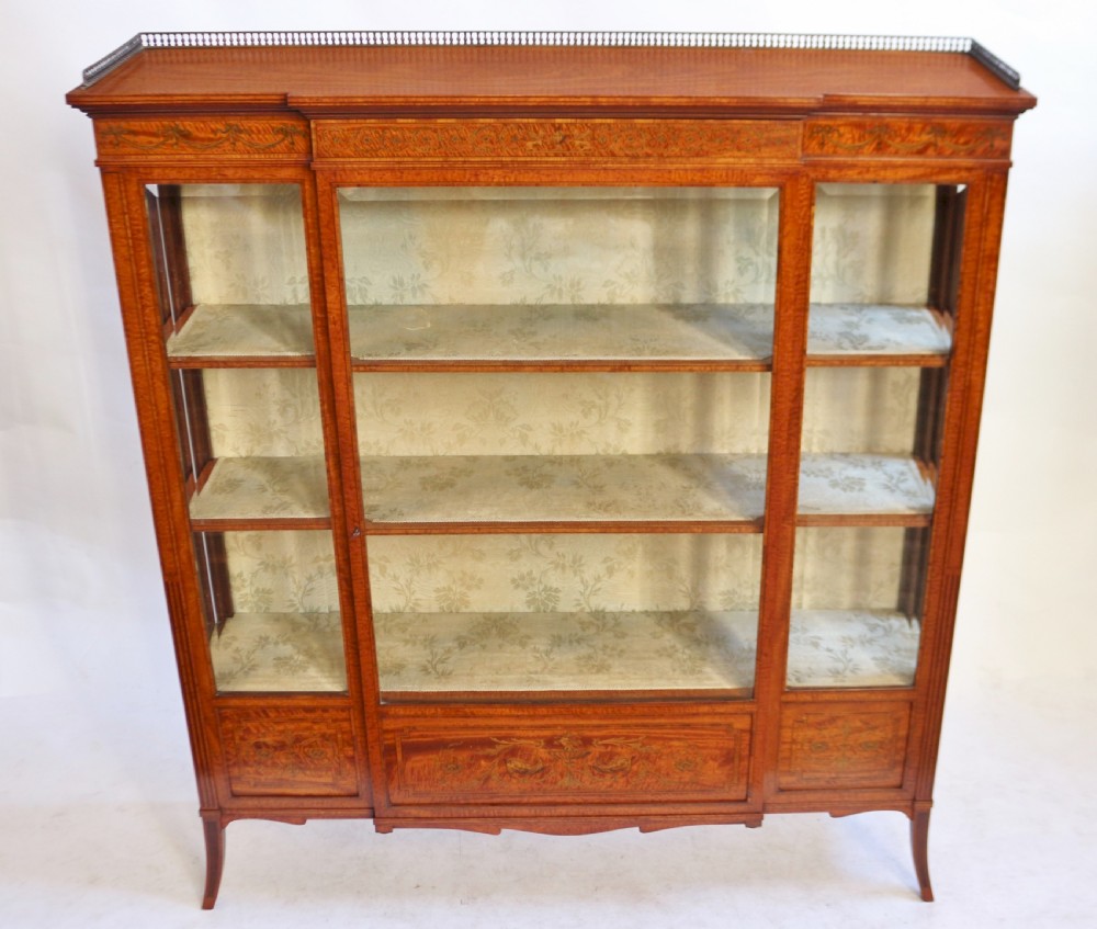 An antique satinwood display cabinet featuring a single central glazed door, fabric-lined interior shelves, floral marquetry inlay, and slightly splayed legs.