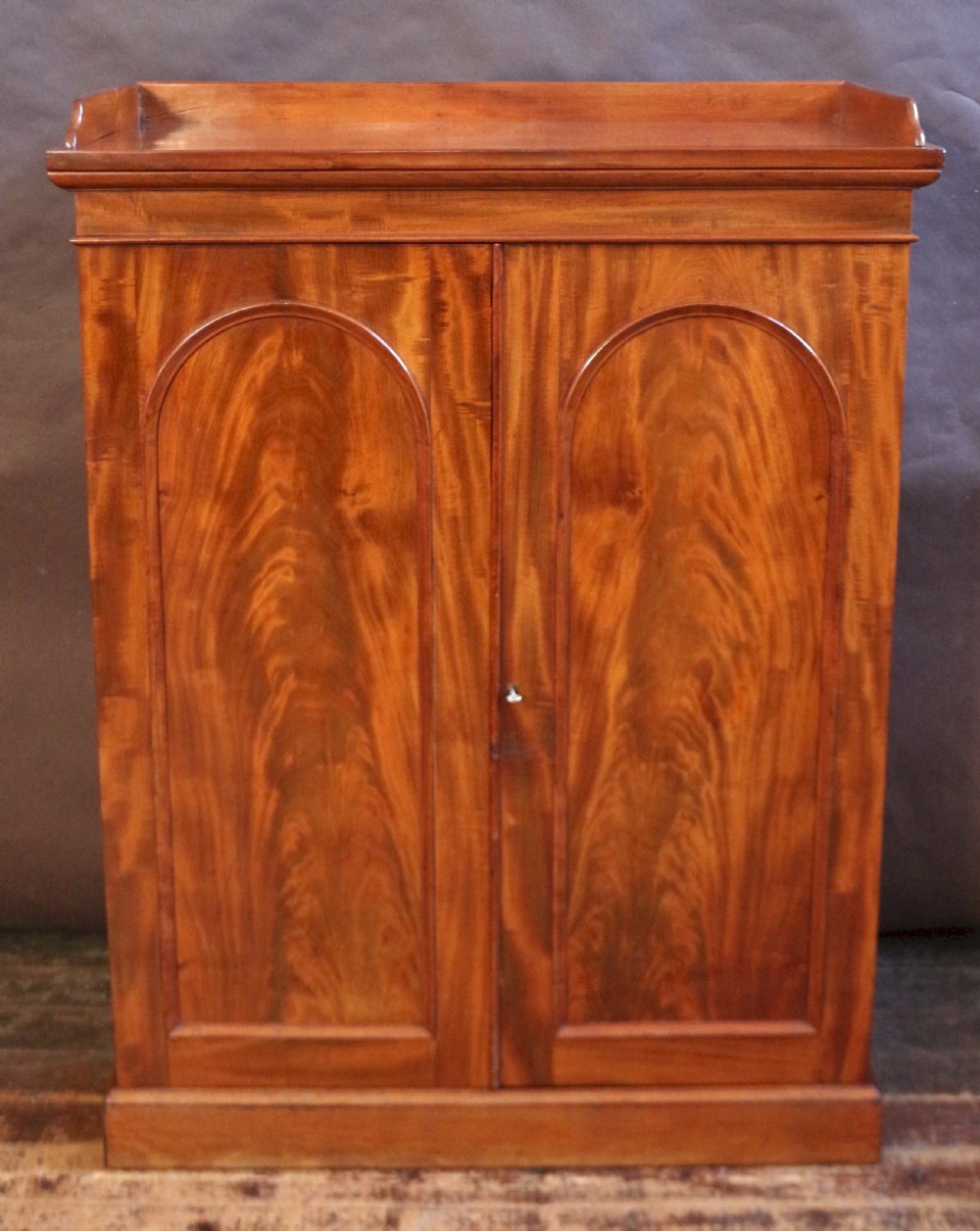 A 19th-century Victorian mahogany two-door cupboard featuring arched panel doors, rich flame veneer patterns, a low rear gallery top, and a solid plinth base.