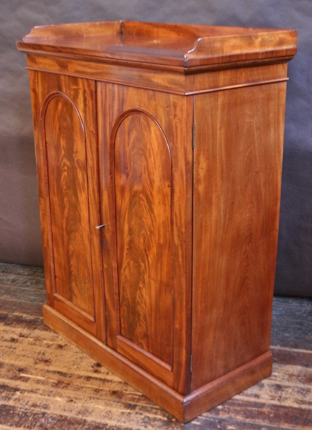 A 19th-century Victorian mahogany two-door cupboard featuring arched panel doors, rich flame veneer patterns, a low rear gallery top, and a solid plinth base. View V