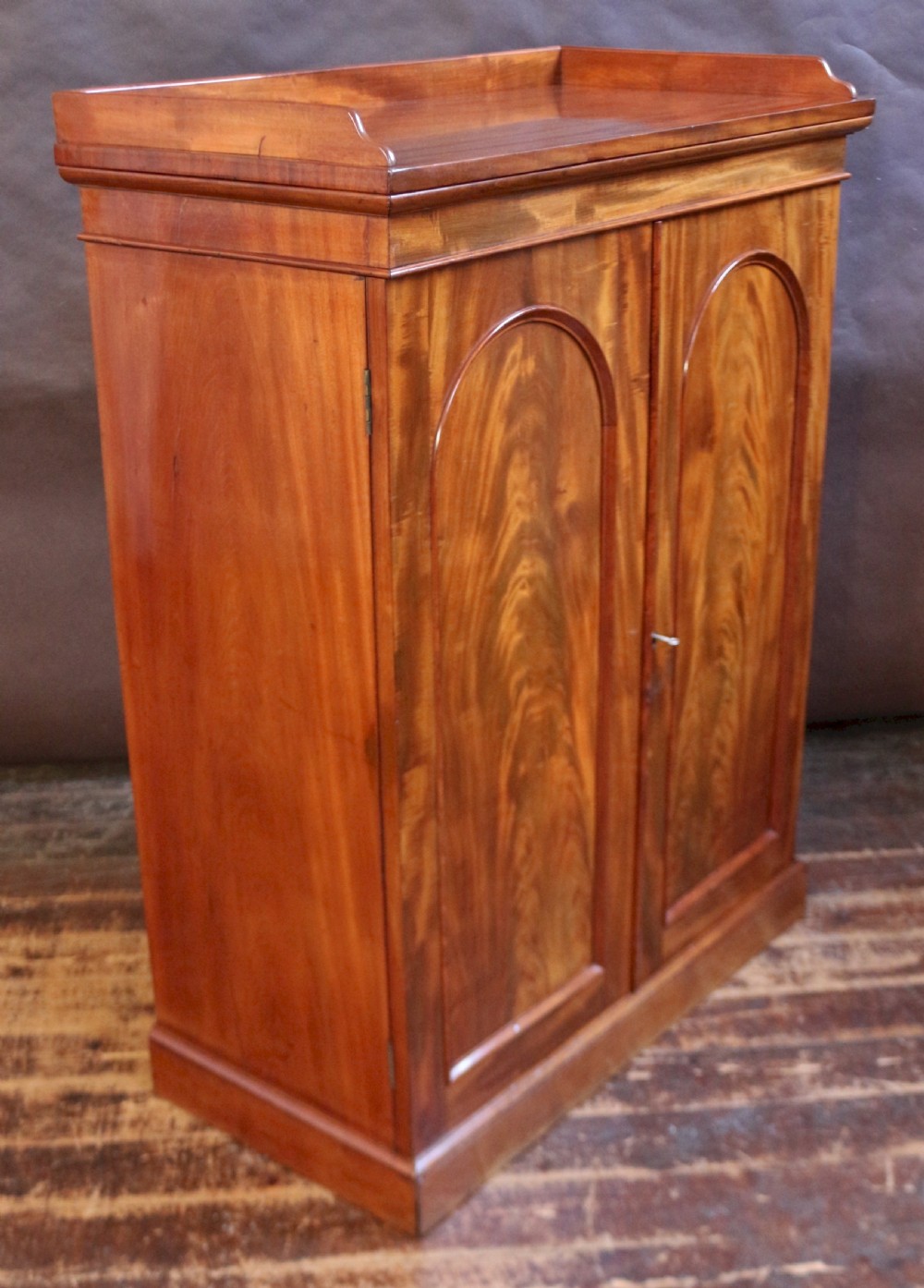 A 19th-century Victorian mahogany two-door cupboard featuring arched panel doors, rich flame veneer patterns, a low rear gallery top, and a solid plinth base. View VI