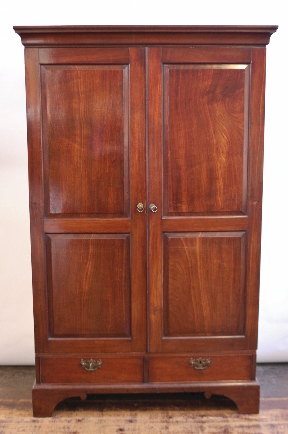 A George III mahogany two-door wardrobe featuring fielded panel doors, two base drawers with brass handles, a moulded cornice, and bracket feet.