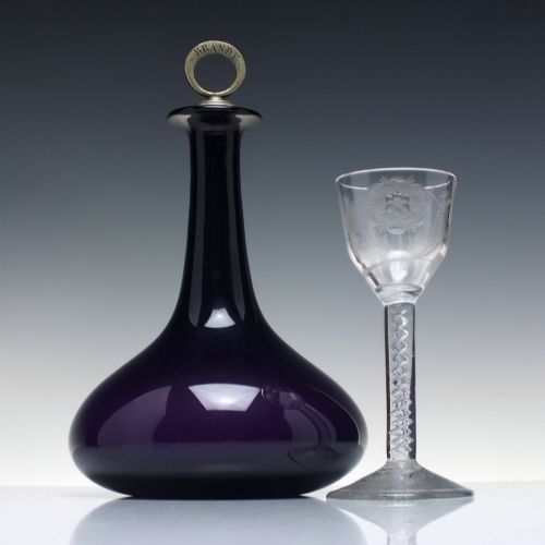 fig 1. A deep purple amethyst glass decanter featuring a wide bulbous base, elongated neck, and a metal ring stopper engraved with the word BRANDY.