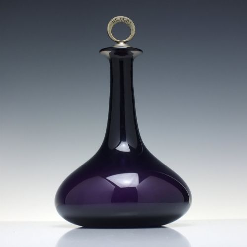 fig 2. A deep purple amethyst glass decanter featuring a wide bulbous base, elongated neck, and a metal ring stopper engraved with the word BRANDY.
