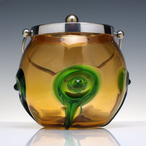 fig 1. An Art Nouveau amber glass biscuit barrel featuring green peacock eye trailing decoration and a silver-plated metal rim, lid, and swing handle.