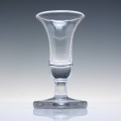 fig 1. An 18th-century clear glass firing glass featuring a flared trumpet rim, short stem element, and a heavy, thick round foot.