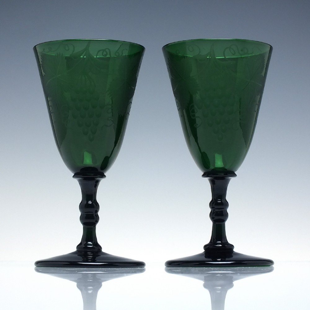 A pair of mid-19th-century dark green glass wine goblets featuring engraved grapevine motifs on the bowls, knopped stems, and circular bases.