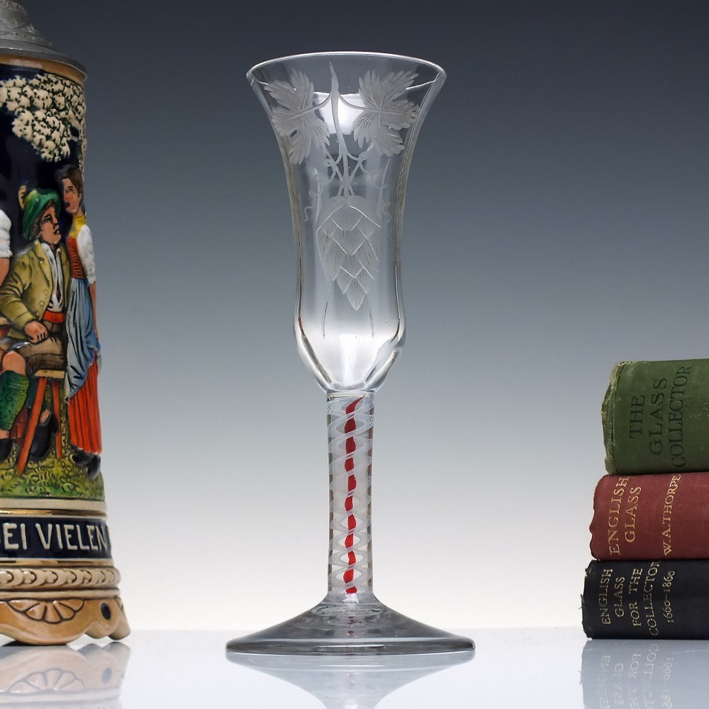 An eighteenth-century clear lead glass ale flute featuring a flared rim, floral hop wheel engravings, and a central stem containing a red and opaque white spiral twist.